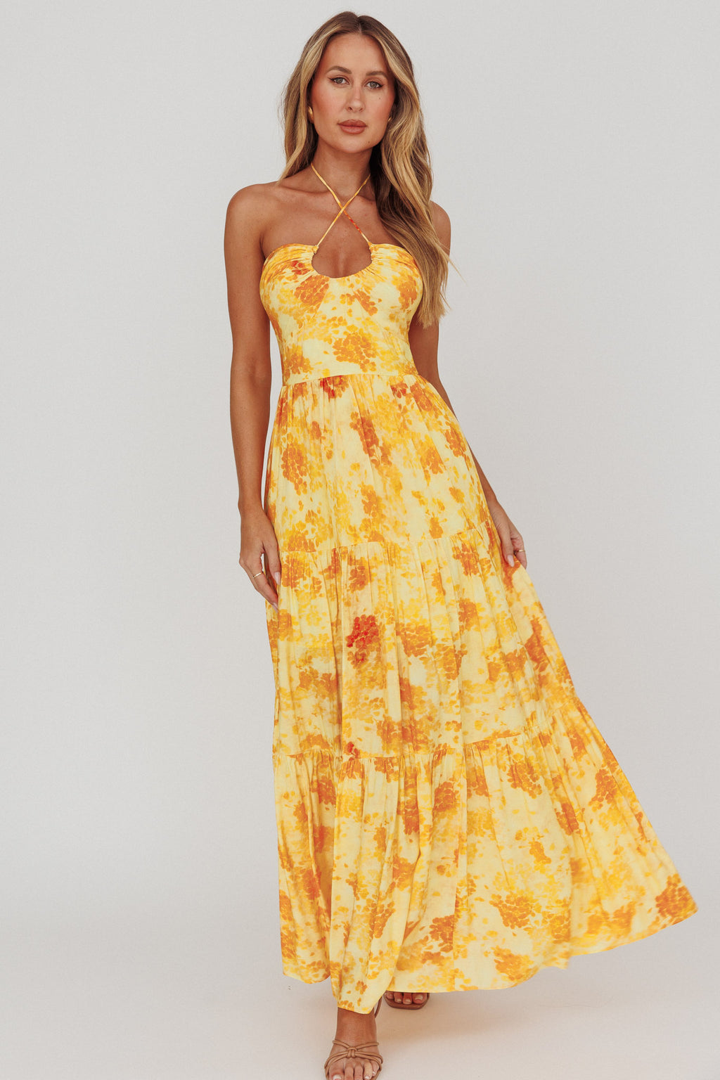 Woman wearing a yellow floral halter neck maxi dress with tiered skirt and front cut-out detail, perfect for summer occasions and wedding guest styling.